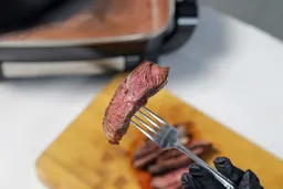 A hand in black glove using a fork to pick up a slice of steak. In the background is the Hamilton Beach Ceramic Non-Stick Electric Skillet 38529K.