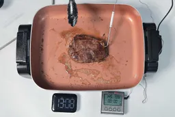 A piece of deep golden brown steak inside the Hamilton Beach Ceramic Non-Stick Electric Skillet 38529K. In front of the skillet is a digital timer and a meat thermometer with its probe inserted into the steak.