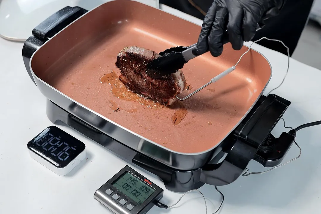 A person holding a steak with a tong to sear the edge of a steak in the Hamilton Beach Ceramic Non-Stick Electric Skillet 38529K. A digital timer, and a meat thermometer.