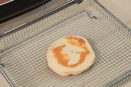 The bottom side of a golden brown pancake inside an air fryer basket. In the corner is the Hamilton Beach Ceramic Non-Stick Electric Skillet 38529K.