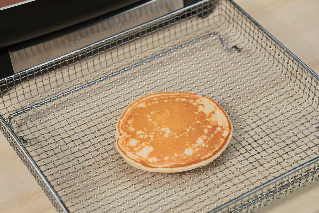 The top side of a golden brown pancake inside an air fryer basket. In the corner is the Hamilton Beach Ceramic Non-Stick Electric Skillet 38529K.
