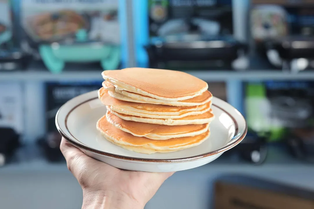 A hand holding a plate of pancakes. In the background is a shelf of electric skillets.