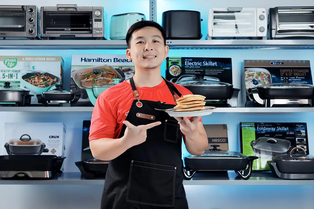 A person holding and pointing at a plate of pancakes. In the background is a shelf of electric skillets and toaster ovens.