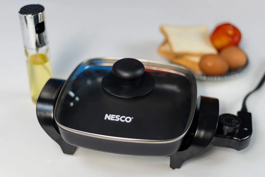 The Nesco 8-inch electric skillet against a blurry white background, with fruit bowl and cooking oil canister in the back.
