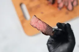A hand in black glove using a fork to pick up a slice of steak. In the background is the Nesco Non-Stick Electric Skillet ES-08.
