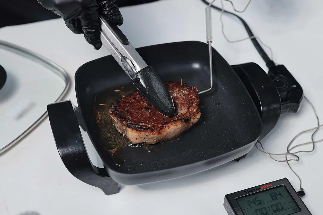 A piece of deep golden brown steak inside the Nesco Non-Stick Electric Skillet ES-08. In front of the skillet is a meat thermometer with its probe inserted into the steak.