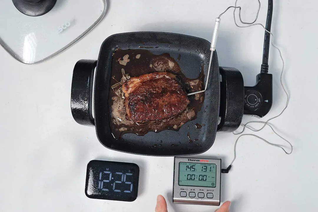 A piece of deep golden brown steak inside the Nesco Non-Stick Electric Skillet ES-08. In front of the skillet is a digital timer and a meat thermometer with its probe inserted into the steak.
