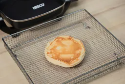 The bottom side of a golden brown pancake that’s dark around the edges inside an air fryer basket. In the corner is the Nesco Non-Stick Electric Skillet ES-08.