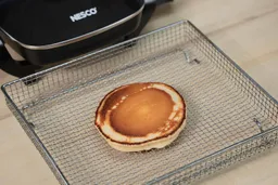The top side of a golden brown pancake that’s dark around the edges inside an air fryer basket. In the corner is the Nesco Non-Stick Electric Skillet ES-08.