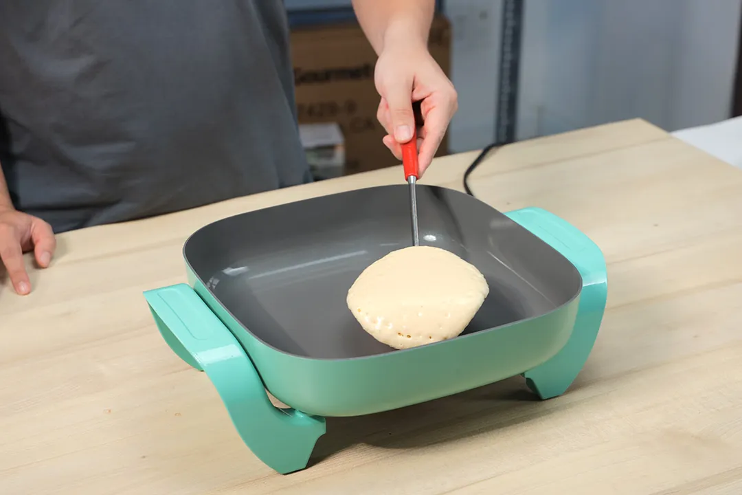 A person flipping a pancake in the GreenLife Ceramic Non-Stick Electric Skillet CC003725-002.