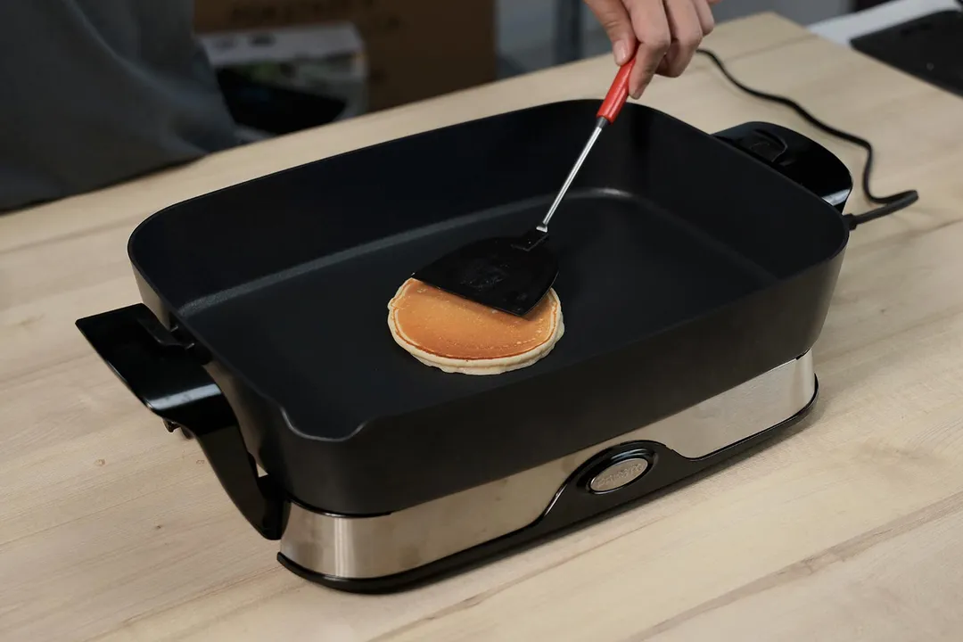 A golden brown pancake inside the Presto Foldaway Electric Skillet 06857. A spatula is pressing down on it.