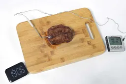A piece of deep golden brown steak with a meat thermometer inserted into it on a wooden cutting board. At the corner is the Presto Foldaway Non-Stick Electric Skillet 06857.