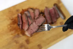 A hand in black glove using a fork to pick up a slice of steak.