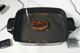 A piece of golden brown steak inside the Presto Foldaway Non-Stick Electric Skillet 06857. In front of the skillet is a digital timer and a meat thermometer with its probe inserted into the steak.