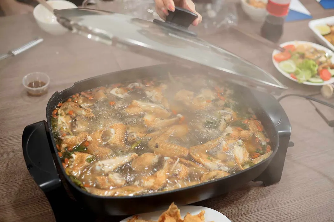 On the dining table, a hand lifts up the lid of the Elite Gourmet Non-Stick Electric Skillet EG-6203# to reveal a steamy chicken hot pot.