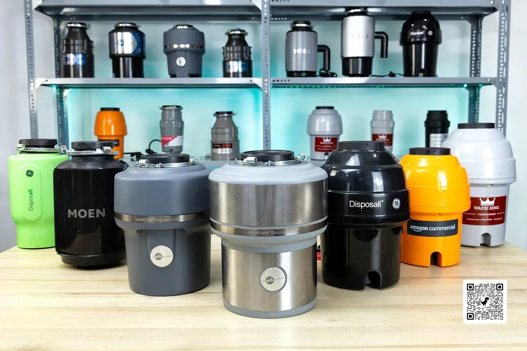 Seven garbage disposal machines on a table, other disposers on a shelf at the back