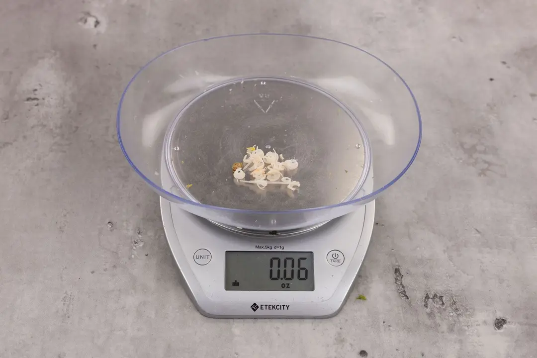 0.06 ounces of ground products from garbage disposal, displayed on digital scale, placed on granite-looking top. Pieces of shredded fish backbone and few pieces of fish bone.