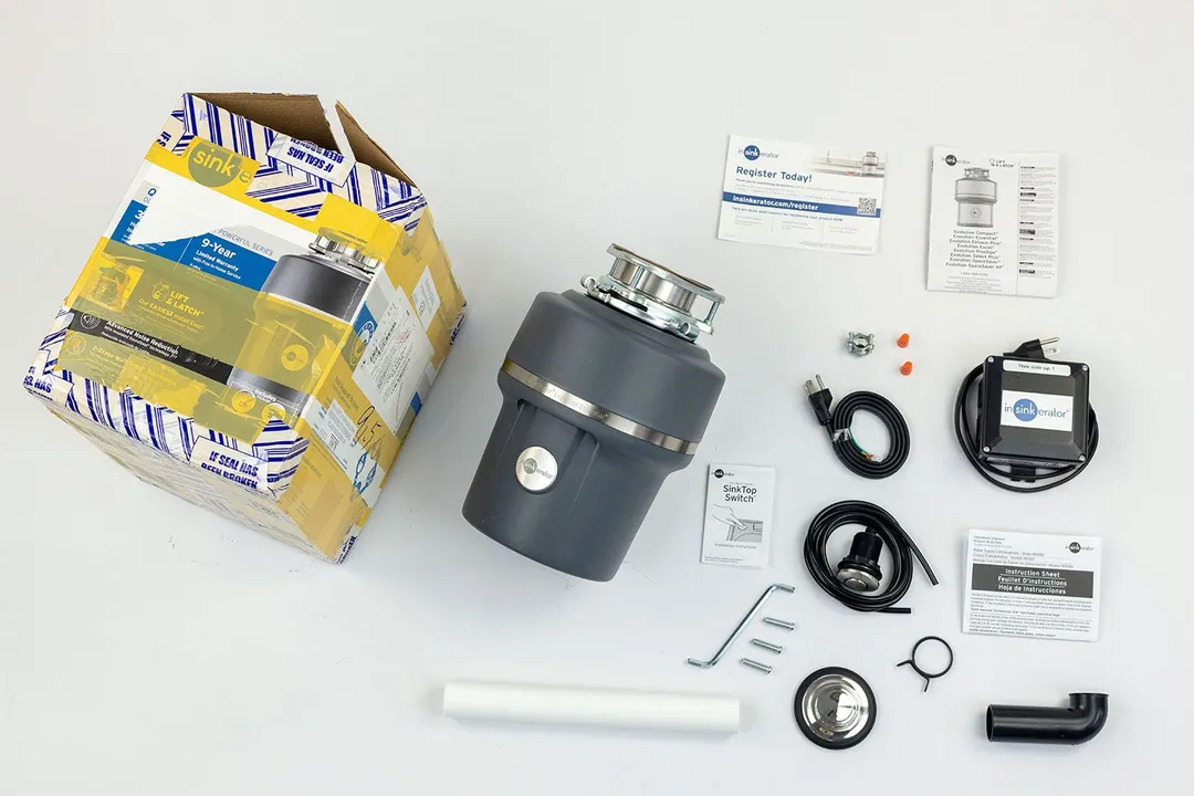 InSinkErator Evolution Essential XTR 3/4 HP with 3-Bolt Mount assembly on top, next to its box, user manual, registration slip, sink stopper, hose clamp, elbow tube, straight tube, wrench, and bolts.