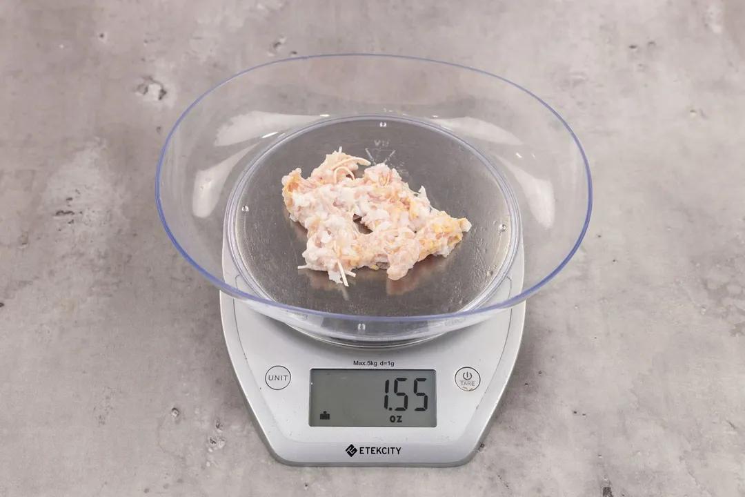 1.55 ounces of ground chicken scraps from garbage disposal, displayed on digital scale, placed on granite-looking table. Visible pieces of shredded tendon and shredded bone among mess of soft tissue.