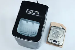 A portable ice maker on a countertop with a glass bowl to the right on a scale with badly formed ice inside weighing 6 grams.