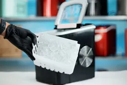 A full basket of ice being taken out of a countertop ice maker with a number of ice makers seen on a shelf in the background.