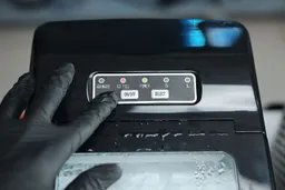 A finger in a black glove pointing to the select button on the control panel of a countertop ice maker while the working indicators are Power in red and Ice Full in green.
