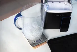 A jug filled with ice bullets in a jug being weighed on a scale showing 330 grams. To the right is a portable ice making machine with a smartphone propped up against the side.