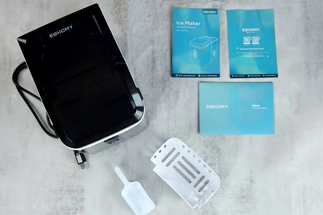 The unboxed EUhomy countertop ice maker with an ice basket and ice scoop in the front of the machine with the user manual, and other bundled