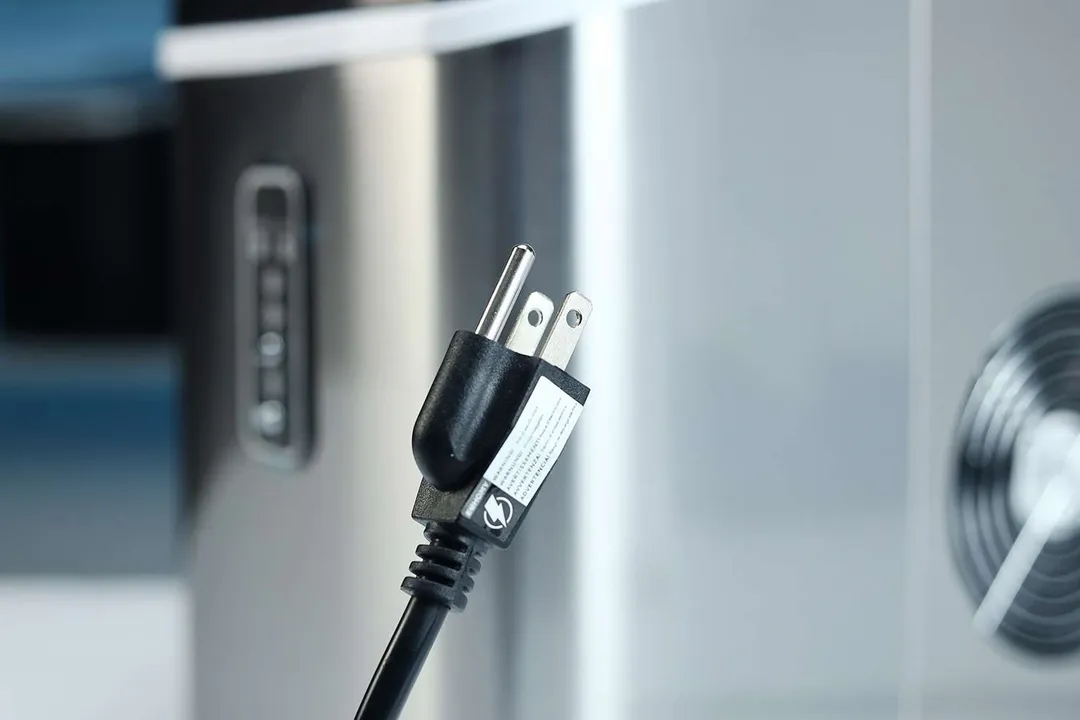 A three-pronged plug at the end of a black cord pictured against the body of a portable ice making machine.