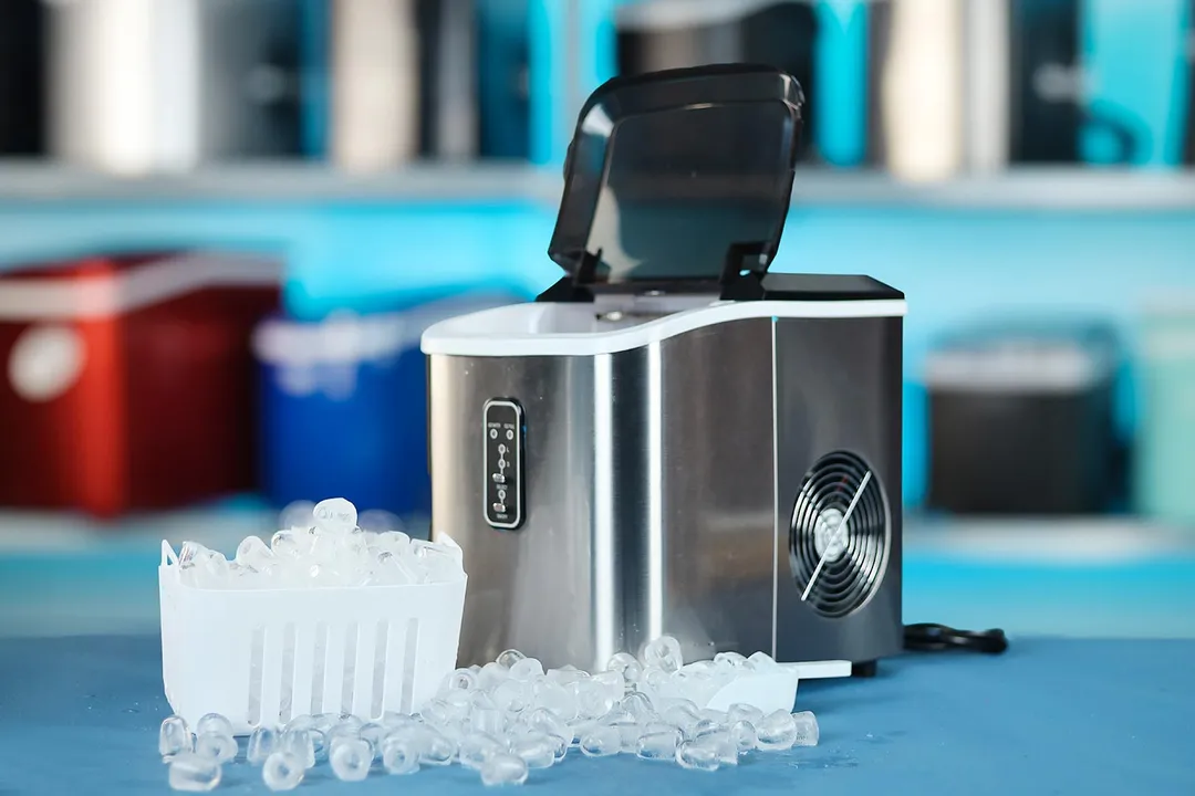 The EUhomy portable ice maker on a countertop with its lid open and a basket of ice in front and ice laying along the side to meet the ice scoop on the right.