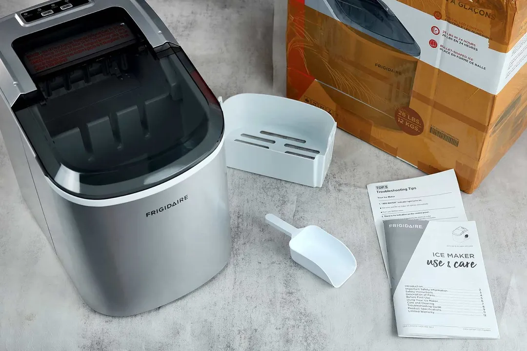 The Frigidaire EFIC189 countertop portable bullet ice maker to the left with its box in the background to the right. In between is the included ice basket, ice scoop, and user manual.