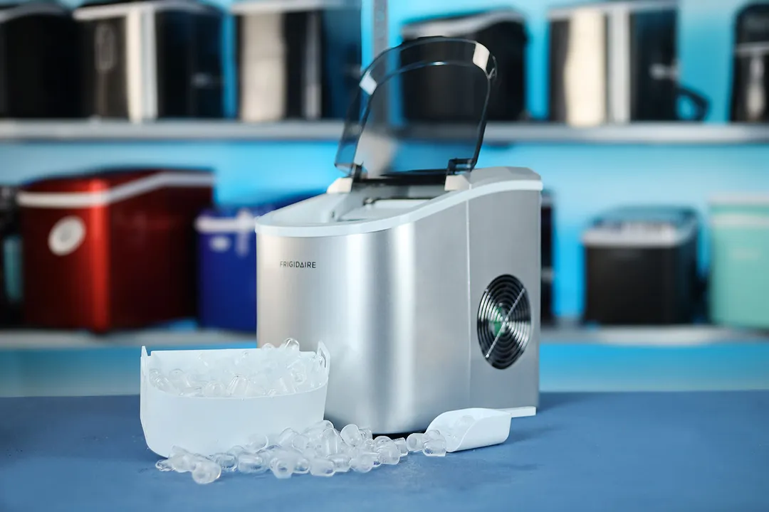 The Frigidaire EFIC189 portable ice maker, lid open on a countertop and a full basket of ice in front with ice scattered around leading to an ice scoop on the right side. 