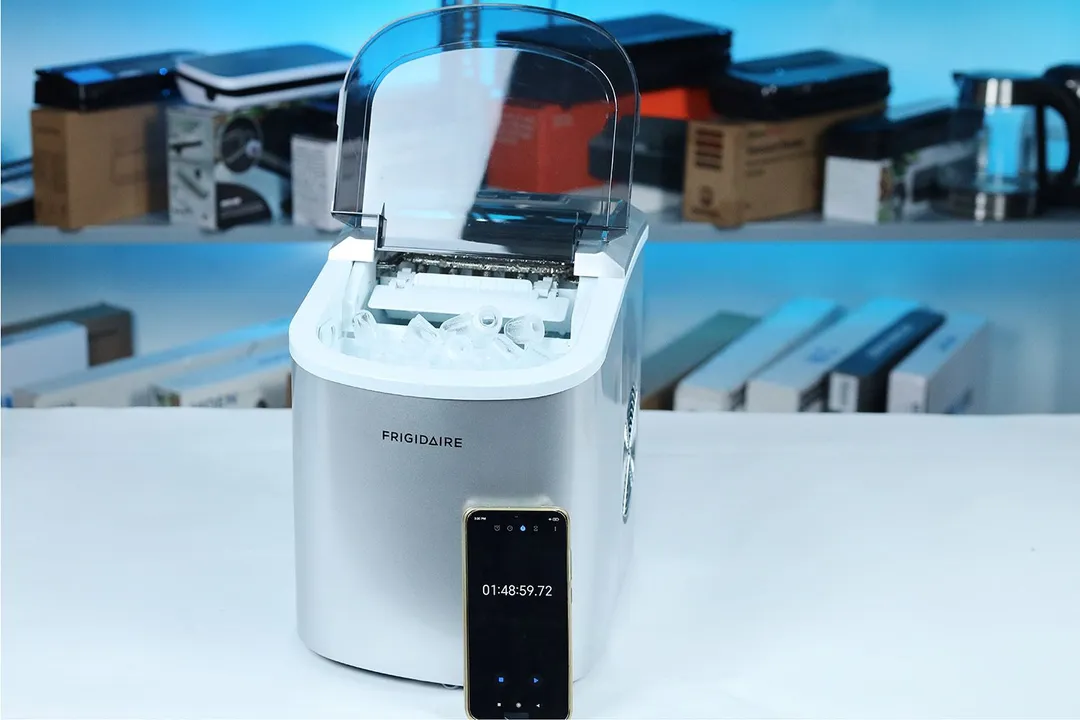 The Frigidaire EFIC189 countertop portable bullet ice maker on a countertop with a full basket of ice and a timer showing 1 hr 48 min. In the background is a shelf with various kitchen appliances.