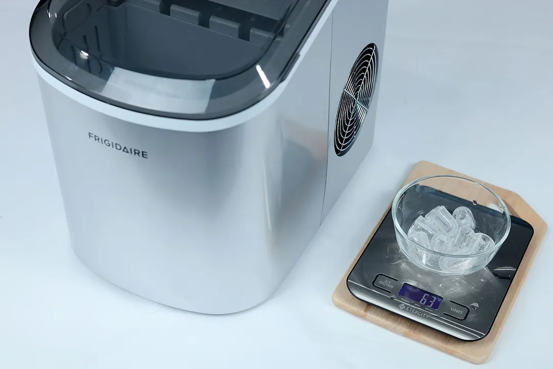 The Frigidaire EFIC189 countertop portable bullet ice maker to the left and a bowl on a scale with one round of nine ice bullets measuring 63 grams.