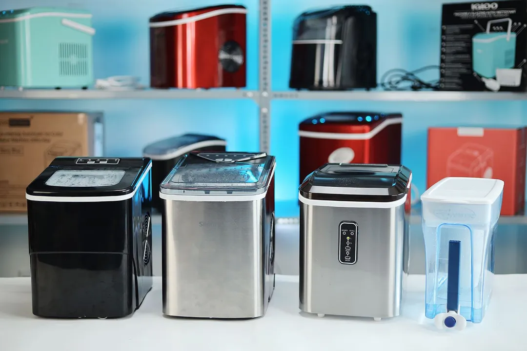 Three portable bullet ice makers on a countertop with a water pitcher filter on the far right. In the background is a shelf with numerous other ice making machines.