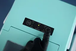 A gloved finger pressing the select button on a countertop ice maker.