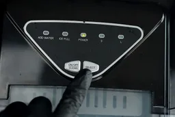 A finger in a black latex glove about to press the Select button on the control panel of a portable countertop ice maker.
