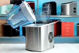 Water from a ZeroWater pitcher is being poured into the reservoir of the Silonn countertop ice maker with a number of other ice makers on a shelf in the background.