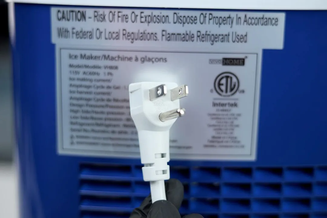 The white three pronged plug of a portable countertop ice maker pictured against the background of the technical information sticker on the back of the machine.
