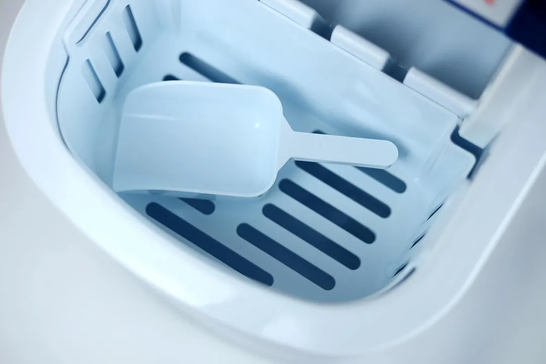 A picture of an empty ice basket resting inside a portable countertop ice making with the scoop placed inside.