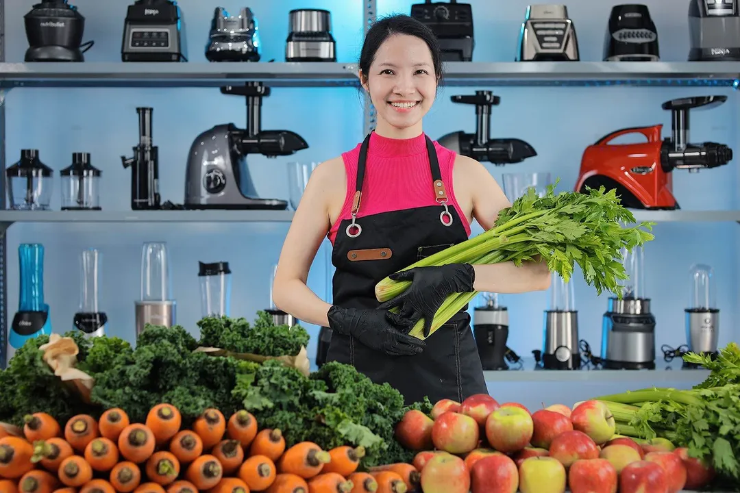 Woman holding celery, with piles of carrots, apples, kale, and celery in front of her