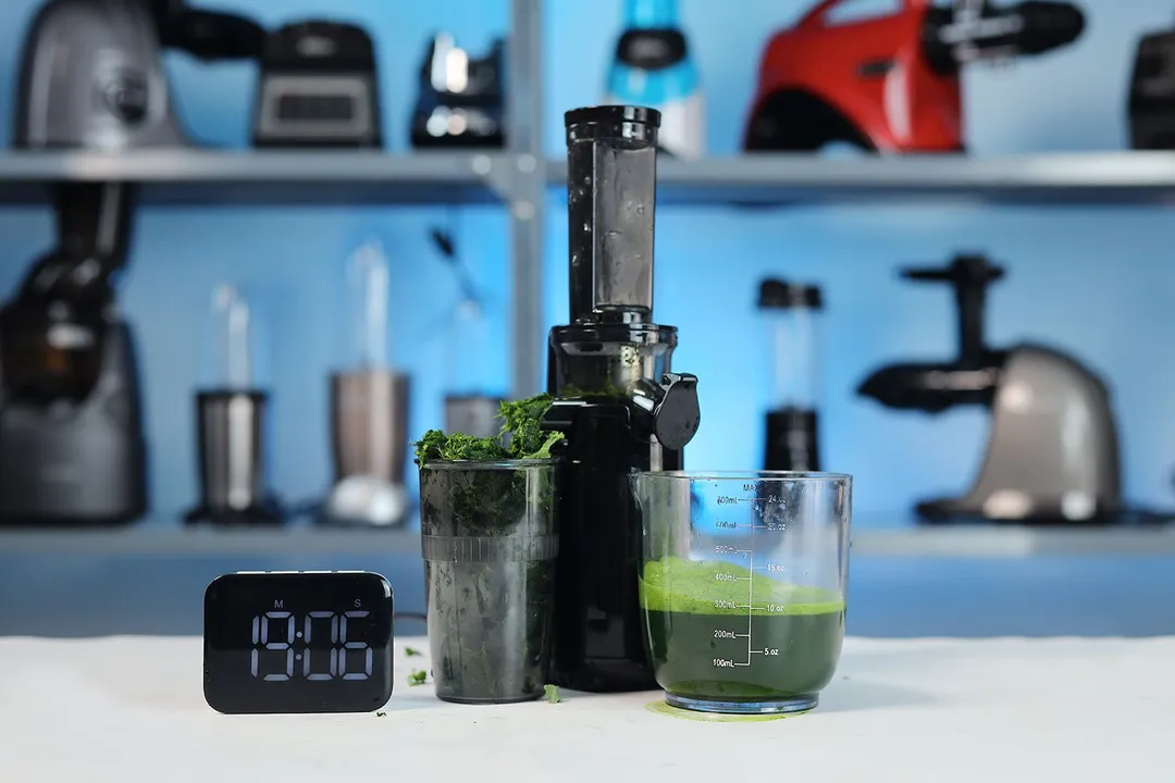 The Elite Gourmet vertical masticating juicer with green juice and pulp in the cups, next to a timer, other juicers and blenders