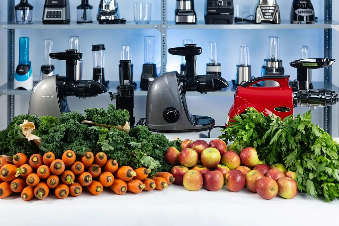 Piles of carrots, kale, apples, and celery on the table with different masticating juicer models