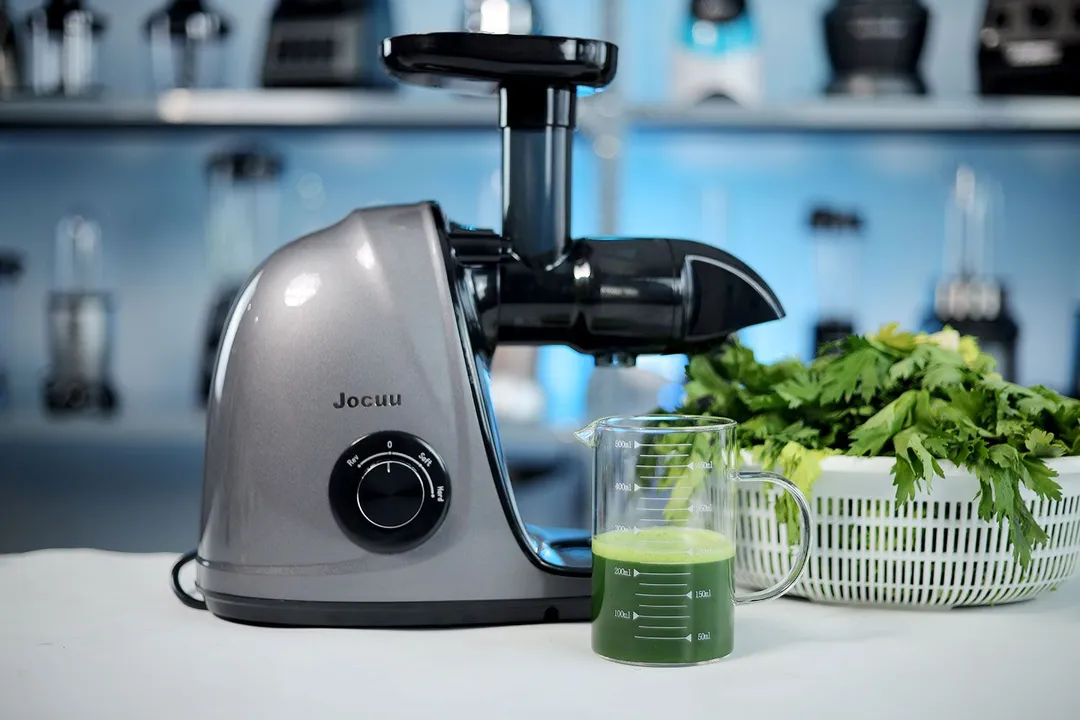 The Jocuu masticating juicer next to a glass of green juice, some celery in a white basket