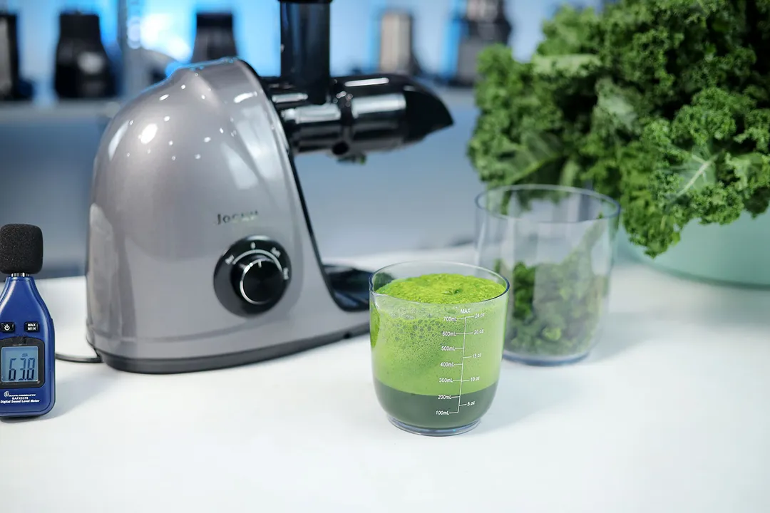 The Jocuu slow juicer after juicing kale, next to a juice cup, pulp container, sound meter, and kale 