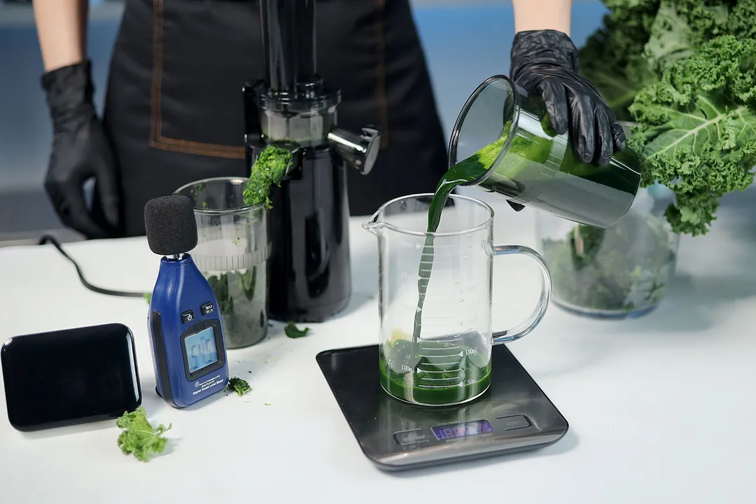 Person pouring kale juice from container into glass on a scale, Elite Gourmet masticator, two baskets of kale, timer, sound meter