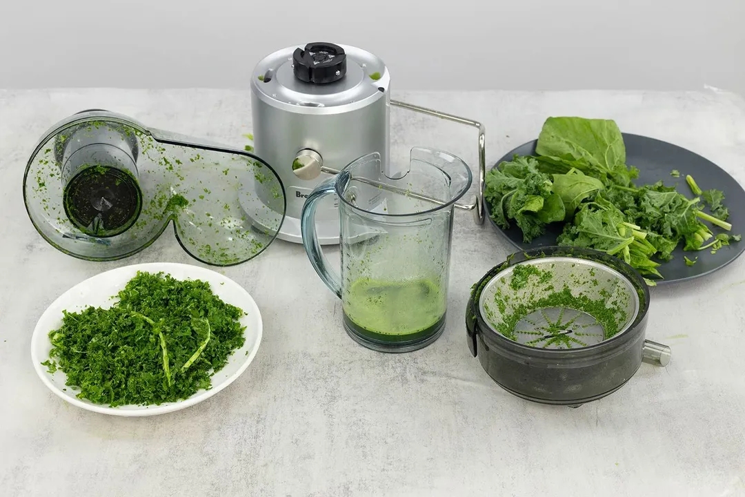 Parts of a centrifugal juicer after juicing leafy greens, next to a plate of green pulp and a plate of green vegetables