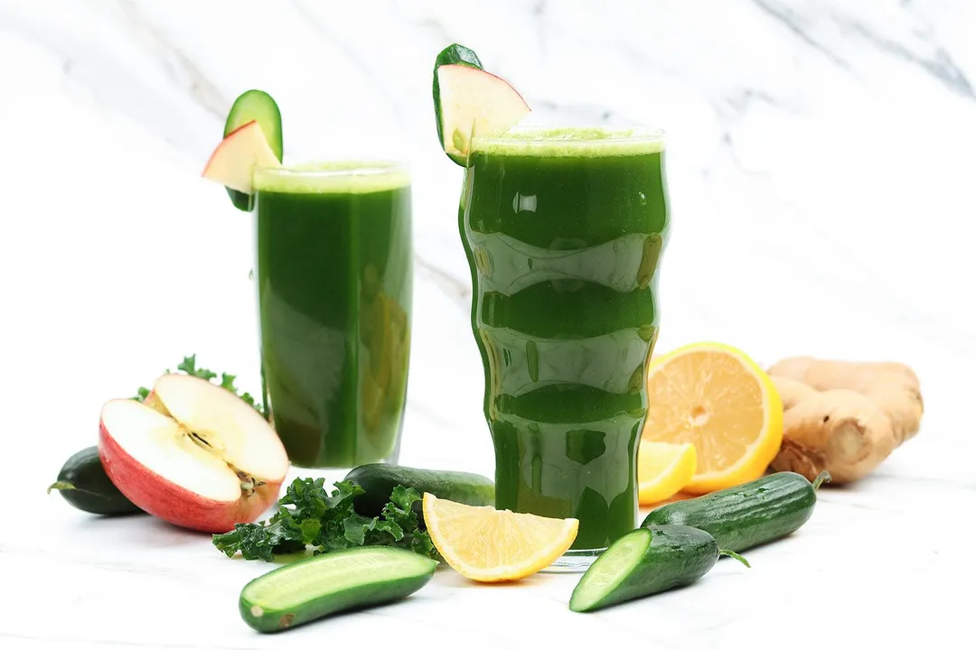 Two glasses of green juice, slices of apple, kale, lemon, and cucumbers