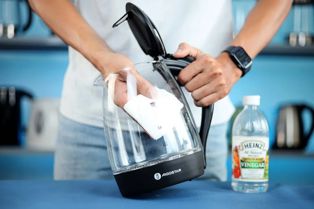 A person in a white shirt holding the Zeppoli Electric Kettle ZPL-KETTLE by its handle on one hand and the other hand wiping the carafe interior with a piece of tissue. On the right is a bottle of white vinegar.