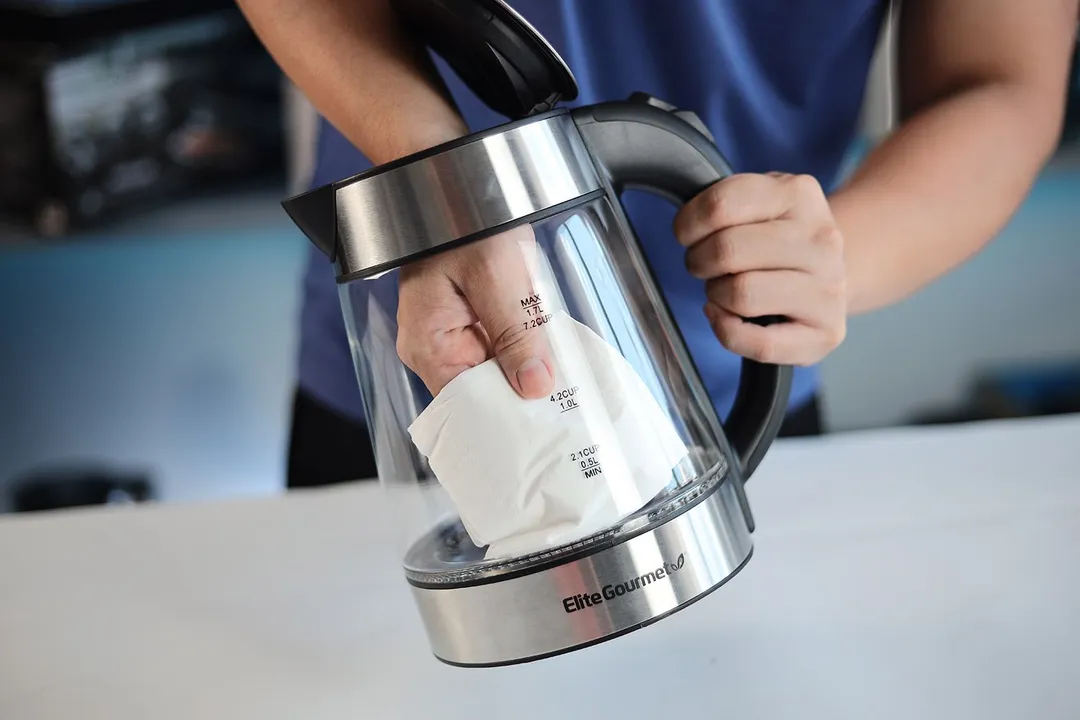 A person in a blue shirt holding the Amazon Basics Electric Glass and Steel Kettle (F-625C) by its handle on one hand and the other hand wiping the carafe interior with a piece of tissue.
